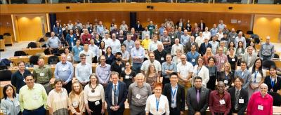 A large group of people pose for a photo in a lecture hall, standing and sitting in rows, with some smiling and wearing conference badges.