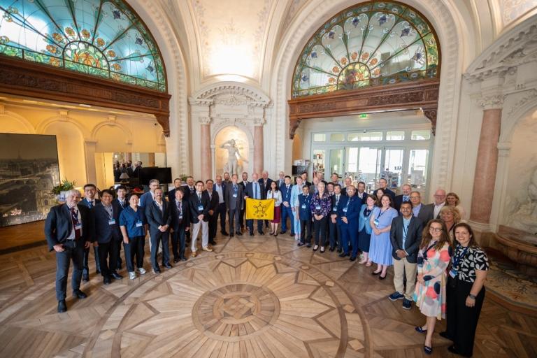 A large group of people in formal and semi-formal attire stands in an ornate room with arched stained-glass windows and a patterned wooden floor.