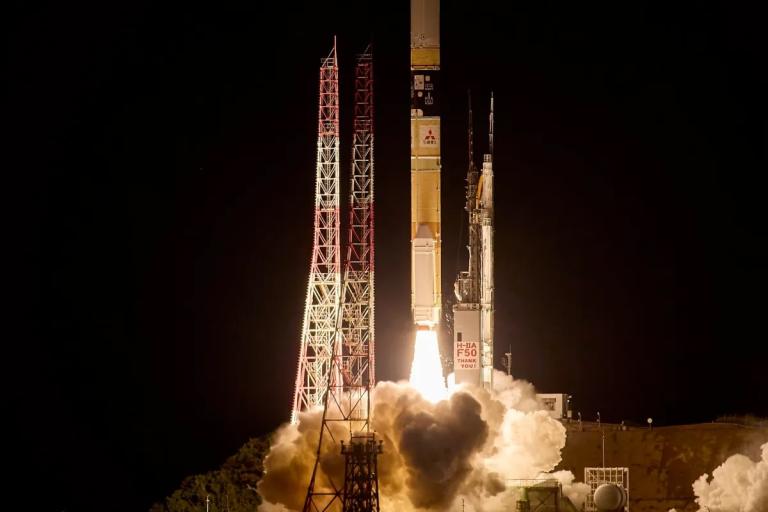 A rocket launches at night, producing bright flames and smoke, with support structures and towers visible beside the launchpad.