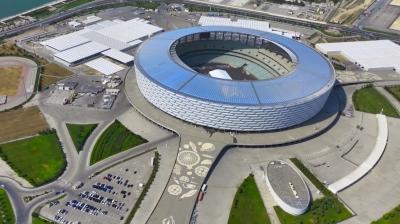 Aerial view of a large, modern stadium with a circular roof, surrounded by parking areas and greenery, located near a body of water.