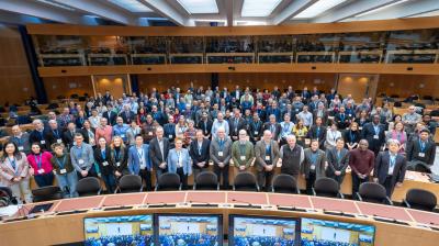 A large group of people posed in a conference room with wooden walls and a curved desk in the foreground. Multiple monitors display a similar view of the room.
