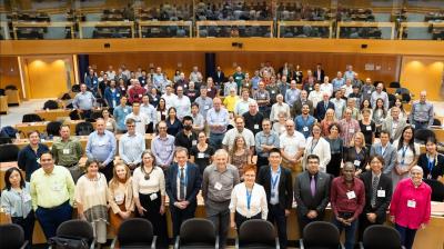 A large group of people pose for a photo in a lecture hall, standing and sitting in rows, with some smiling and wearing conference badges.