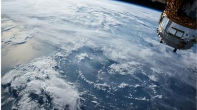 View of Earth from space with a satellite in the top right corner, showing clouds over a large body of water.