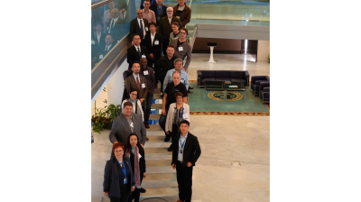 A group of people in business attire pose for a photo on an indoor staircase in a modern building with glass railings.