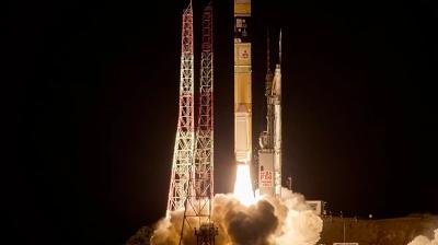 A rocket launches at night, producing bright flames and smoke, with support structures and towers visible beside the launchpad.