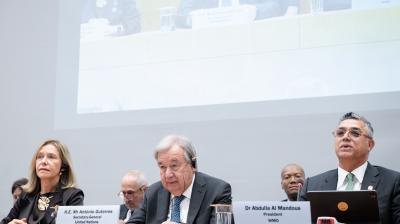 Three officials sit at a conference table with microphones and nameplates; a projected image of the panel is shown above them.