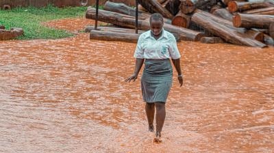 A student in uniform walks barefoot through a flooded, muddy area during heavy rain, with stacked logs in the background.