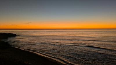 Calm ocean waves under a clear sky at sunset, with an orange glow on the horizon and silhouettes of land on the left side.