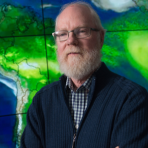 A man with white hair and beard, wearing glasses and a dark sweater, stands with arms crossed in front of a large screen displaying a map with green areas.