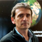 A man with dark hair and a serious expression is in front of a background featuring a partial view of Earth. He is wearing a dark sweater over a light blue collared shirt.