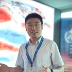 A man wearing a white shirt and lanyard speaks in front of a screen showing a weather map. The background includes a banner with the World Meteorological Organization logo.